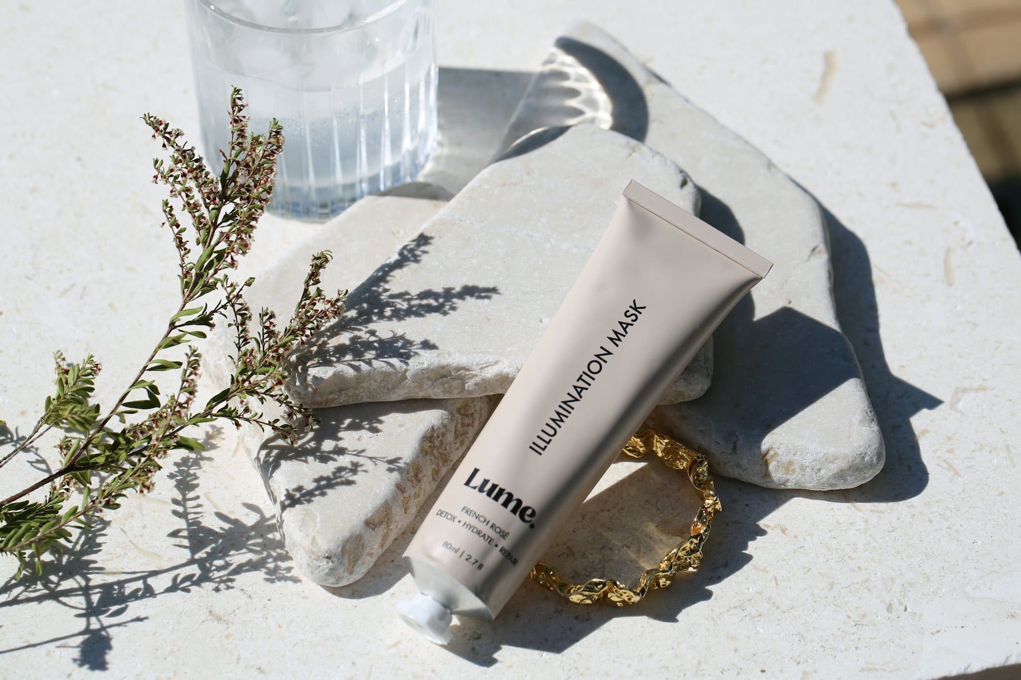 A skincare tube resting on pale stone beside a sprig of dried herb